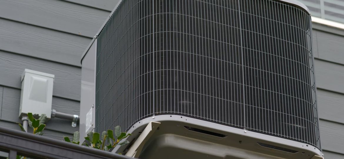 A modern outdoor air conditioning condenser unit mounted on a bracket against the grey siding of a house.