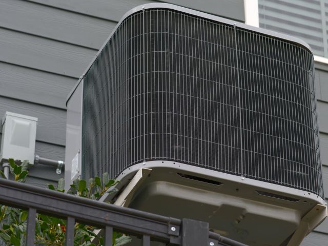 A modern outdoor air conditioning condenser unit mounted on a bracket against the grey siding of a house.